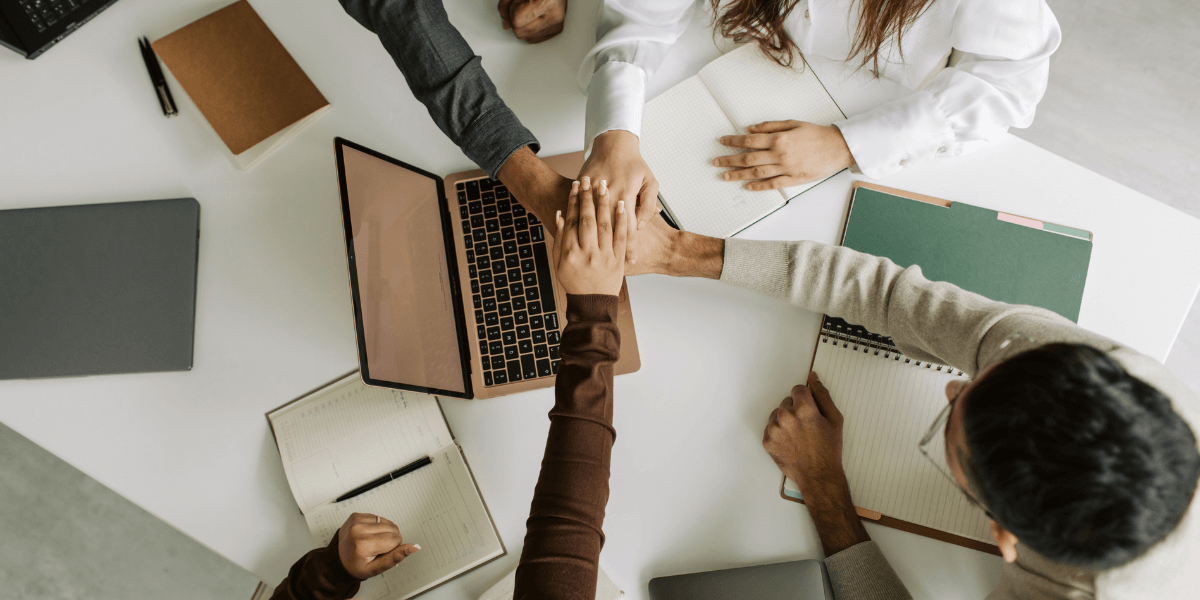 A team of four sales members meeting with their hands all centered over a laptop in the middle of the table.