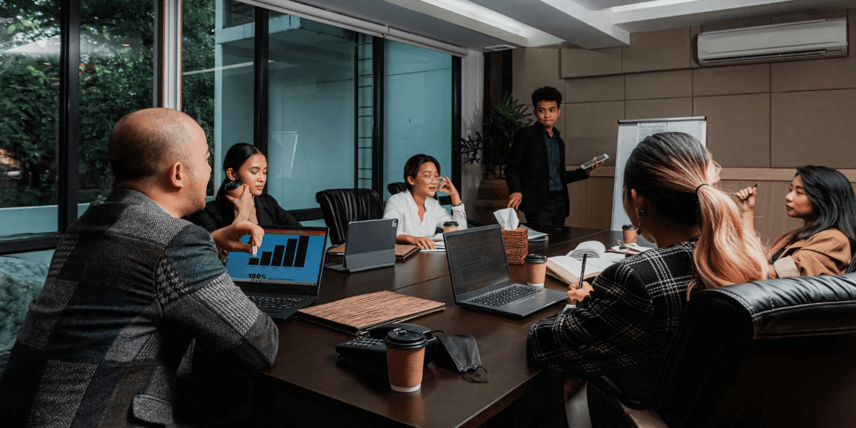 Sales team meeting around a conference table with laptops, one showing a bar chart, as a colleague presents at a whiteboard