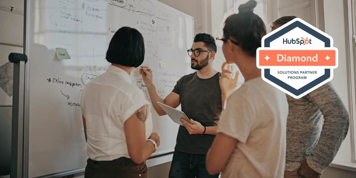 A group of marketers looking at a white board while discussing marketing techniques. there is a HubSpot Diamond partner solutions badge overlayed to the right representing Vested Marketing's HubSpot partnership