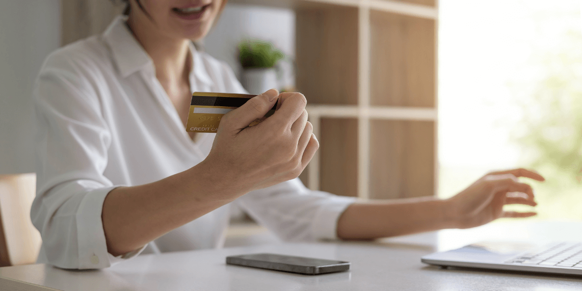 A woman in a white shirt who is using a credit card to pay for something while on a laptop, sitting at a table with a phone on the table in front of her.