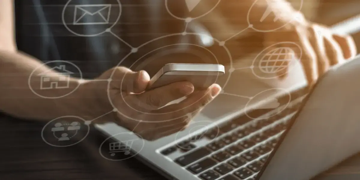 Person holding a smartphone and typing on a laptop with digital icons for email, shopping, and cloud services overlayed, symbolizing digital connectivity and online business.