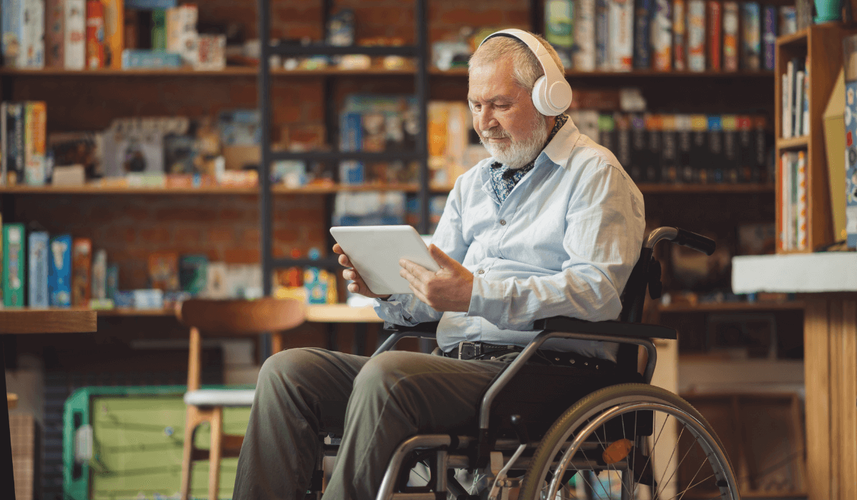 Man in a wheelchair using a tablet with headphones on in a library. 