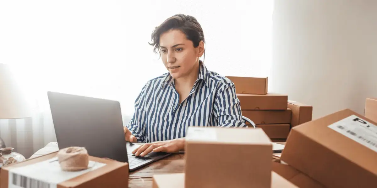 A woman surrounded by cardboard boxes while looking at her laptop and taking inventory.